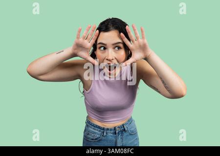 Giovane donna che esprime sorpresa con le mani alzate e la bocca aperta, eccitata e stupita su uno sfondo verde Foto Stock