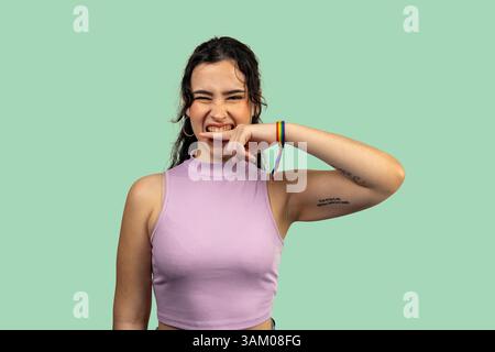 Ritratto in studio di una giovane donna allegra con tatuaggi e un braccialetto arcobaleno che le mordono il dito e si strizzano contro un vibrante backgroun color verde acqua Foto Stock