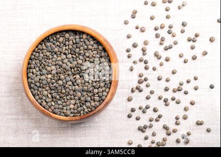 Le lenticchie Puy in una ciotola di legno su lino. Varietà secca di piccole lenticchie, a chiazze, di colore grigio ardesia a verde scuro, con marmorizzazione blu. Esculenta della lente. Foto Stock