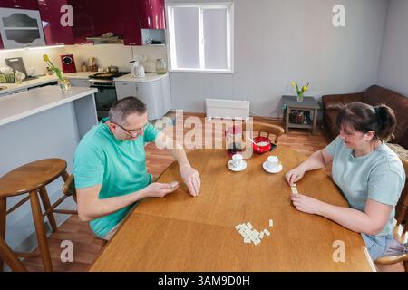 Adulti maturi sono seduti a un tavolo impegnati in un gioco di domino in casa con illuminazione luminosa e altri mobili intorno. Foto Stock