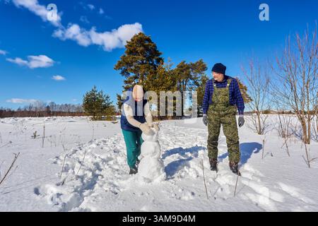 Una coppia di mezza età forma insieme un pupazzo di neve condividendo momenti di gioia all'aperto durante una passeggiata sul bordo della foresta circondata da neve brillante e cielo limpido. Foto Stock