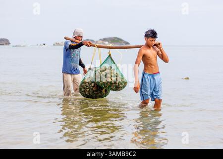 Due pescatori trasportano un sacco di conchiglie raccolte dalla loro barca mentre guado in profondità nel mare a Isla Gigantes Iloilo Filippine Foto Stock
