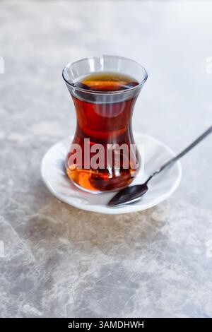 Vetro a forma di tulipani riempito di tè turco caldo Chai poggia su un piattino bianco con un cucchiaio su un tavolo di marmo grigio Foto Stock