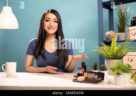 Allegro influencer di bellezza che presenta un tutorial sul trucco, creando contenuti per il suo canale di vlogging. La donna asiatica gesta con la mano mentre spiega lo scopo del suo video durante la trasmissione in diretta. Foto Stock
