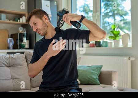 L'uomo applica una pistola massaggiante per alleviare la tensione nella zona del collo e delle spalle. Atleta maschile che esegue la terapia di auto-massaggio con dispositivo a percussione per il recupero dopo l'allenamento Foto Stock
