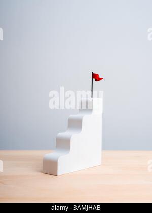 Bandiera rossa vincente in cima alla scala bianca, mentre il grafico a barre si posiziona su un tavolo in legno, sfondo verticale bianco. Processo di crescita aziendale, profitto e investimenti Foto Stock