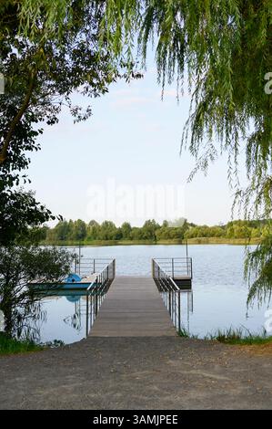 Molo in legno che conduce all'acqua, con catamarano ormeggiato, con ringhiere in metallo sulla riva del pittoresco stagno, fiume, lago, circondato da alberi. Concetto di vacanza estiva in acqua, ricreazione in campagna Foto Stock