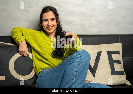 Donna sorridente che punta il telecomando verso la televisione, guarda il suo programma tv o film preferito, rilassati sul comodo divano di casa Foto Stock