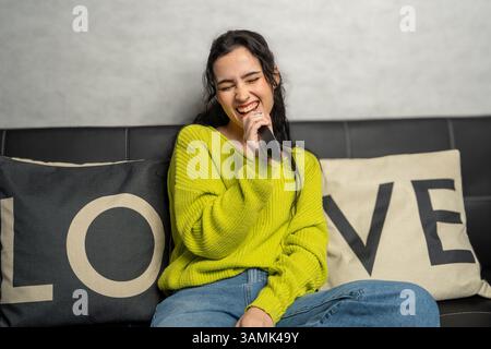 Giovane donna felice che canta karaoke, tiene il telecomando come microfono, siede sul divano con cuscini d'amore Foto Stock