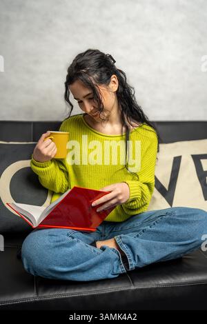 Giovane donna che si gode un momento di relax leggendo un libro e bevendo un caffè su un comodo divano Foto Stock