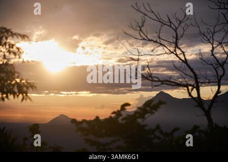 lo splendido tramonto si dispiega dietro una catena montuosa, diffondendo calde sfumature nel cielo. Sagome di alberi incorniciano la scena, esaltando l'atmosfera tranquilla Foto Stock