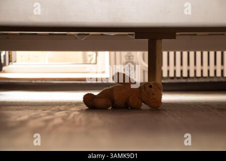 Un orsacchiotto marrone abbandonato giace sotto il letto nella camera da letto di casa, un orsacchiotto sotto il letto Foto Stock