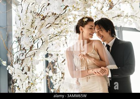 Gli sposi e gli sposi gioiosi condividono un momento intimo durante il loro matrimonio, abbracciato da fiori bianchi. Foto Stock