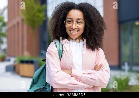 Una giovane donna sorridente con capelli ricci posa all'aperto davanti a un edificio con uno zaino sulle spalle, irradiando positività. Foto Stock