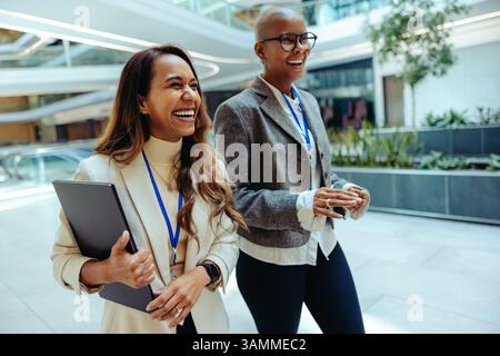 Due allegre donne professionisti che camminano all'interno in un ambiente aziendale. Sono impegnati nella conversazione, indossando sia abiti da lavoro che lanya Foto Stock