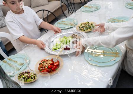 I bambini posizionano con cura un tradizionale piatto di sidro pasquale su un tavolo da pranzo festivo, circondato da cibi simbolici ed eleganti stoviglie natalizie. Foto Stock