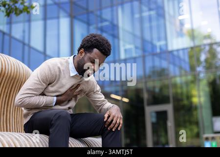 Un uomo fa soffrire il petto mentre è seduto all'aperto vicino a un moderno edificio in vetro. L'immagine suggerisce un problema di salute o disagio. Foto Stock
