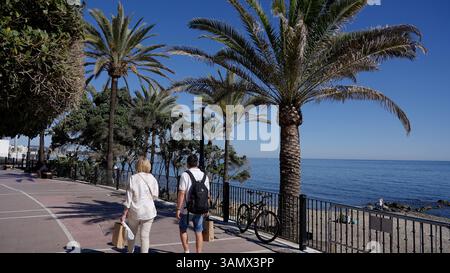 Coppia senior che cammina su una passeggiata soleggiata con palme lungo il Mar Mediterraneo a Marbella, Spagna - MARBELLA, SPAGNA - 30 MARZO 2025 Foto Stock