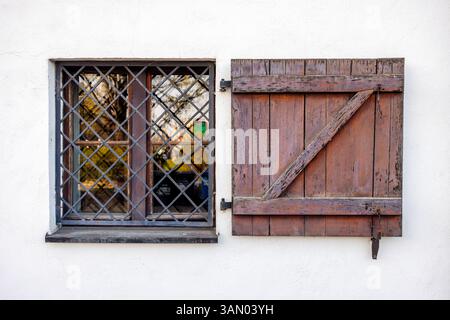 Edificio agricolo europeo con piccola finestra barrata con tapparella in legno inclinabile e pareti intonacate bianche. Barre sulla finestra, nessuna persona, spazio per copiare. Foto Stock