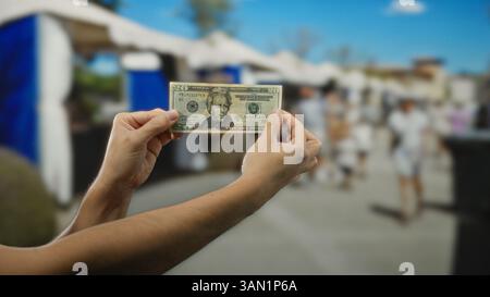 Uomo che tiene in mano un dollaro americano in una strada all'aperto che espone transazioni finanziarie sullo sfondo di un negozio affollato. Foto Stock