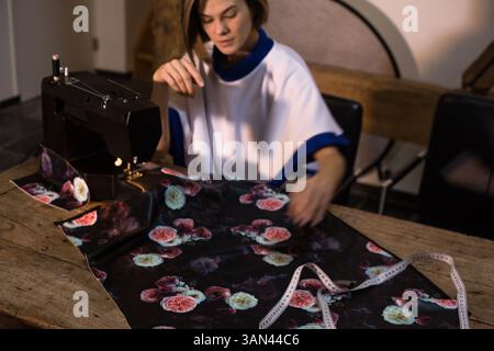 Giovane donna designer che cucisce il tessuto floreale nero su una macchina da cucire per la casa nera su un tavolo di legno. In movimento , flusso di lavoro. Foto Stock