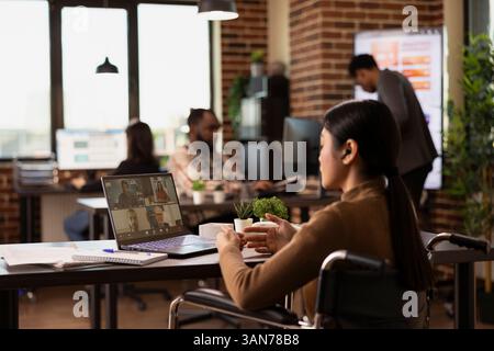 Una donna dirigente d'azienda in sedia a rotelle partecipa a una videoconferenza con potenziali investitori dalla sua scrivania. La manager asiatica tiene una discussione virtuale con i suoi colleghi remoti. Foto Stock