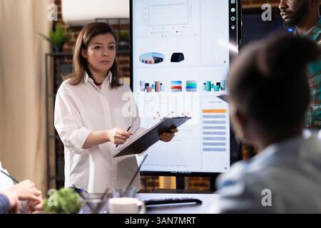Una manager caucasica con appunti presenta le strategie di progetto ai colleghi seduti, utilizzando grafici finanziari e dispositivi digitali per la pianificazione aziendale. Una donna professionale condivide idee di ricerca Foto Stock