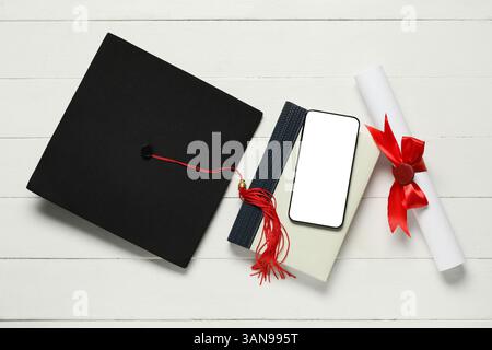 Cappello di laurea, libro, telefono cellulare e diploma su sfondo bianco in legno, vista dall'alto Foto Stock