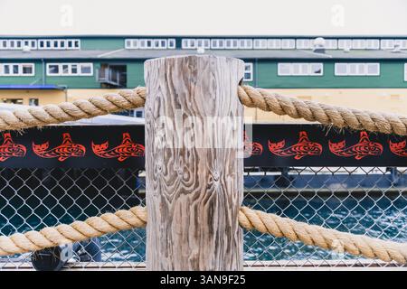 Un palo di legno con una corda legata ad esso e un design nero e rosso su di esso. La recinzione è dietro il palo e il palo è vicino a un corpo d'acqua Foto Stock
