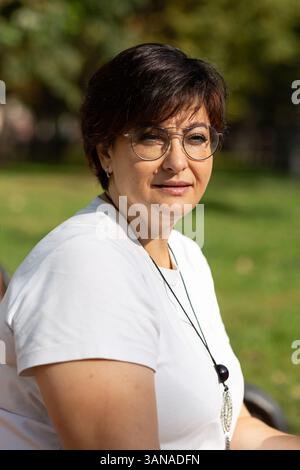 Una bella donna di mezza età con occhiali e capelli scuri corti è seduta su una panchina Foto Stock