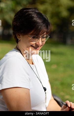 Una bella donna di mezza età con occhiali e capelli scuri corti è seduta su una panchina Foto Stock