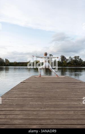 Una donna matura che pratica yoga, Virabhadrasana, posa guerriera, allenamento, vestita con abiti sportivi. sfondo naturale all'aperto. Serata autunnale al ri Foto Stock