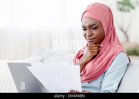 Donna musulmana nera premurosa che controlla i documenti e lavora sul computer portatile a casa Foto Stock