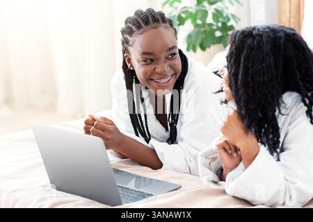 Fidanzate nere positive che si stendano sul letto a casa, usando il laptop Foto Stock