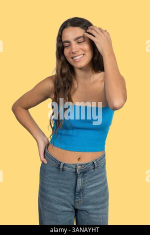 Giovane donna alla moda che si tocca i capelli e sorride su un vivace sfondo giallo Foto Stock