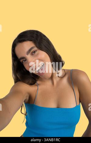 Una giovane donna con lunghi capelli marroni che indossa la parte superiore blu scatta un selfie mentre ride su uno sfondo giallo Foto Stock