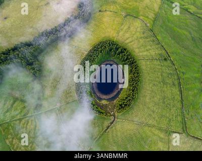 Vista aerea di Lagoa Pau Pique circondata da lussureggianti foreste e campi panoramici, Capelas, Portogallo. Foto Stock