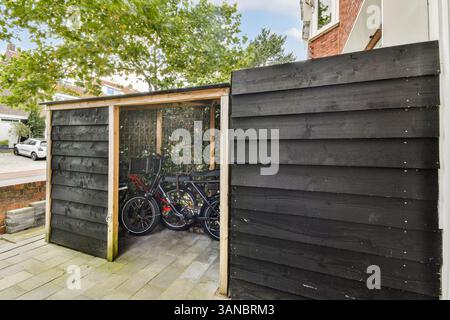 Un elegante capannone per biciclette realizzato in tavole di legno scuro, situato in un quartiere periferico con vegetazione sullo sfondo. Foto Stock