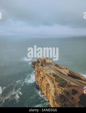 Vista aerea delle scogliere spettacolari e un faro che si affaccia sull'oceano al tramonto, Nazare, Portogallo. Foto Stock