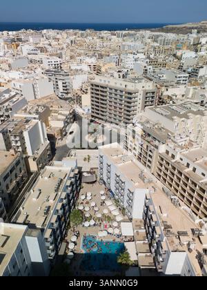 Vista aerea di una vivace festa in piscina con persone che si godono la giornata di sole circondate da edifici residenziali e dal mare, la baia di St. Paul, Malta. Foto Stock