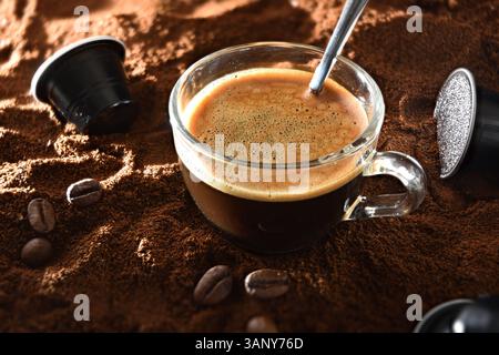 Dettaglio di una tazza di vetro con caffè espresso su caffè macinato con capsule e chicchi di caffè. Vista dall'alto in alto. Foto Stock
