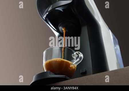 Macchina per caffè a capsule per preparare l'espresso in una tazza di vetro su un banco da cucina in legno con sfondo marrone isolato. Vista dal basso. Foto Stock