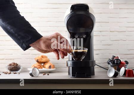 Raccogliere a mano una tazza di caffè appena estratto da una macchina per espresso a capsule su una panca di legno con capsule di caffè, chicchi, pasticcini e un po' di caffè Foto Stock