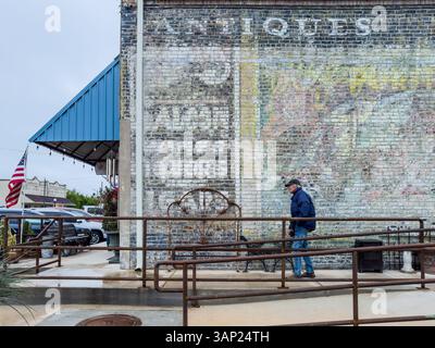 Uomo dei 80 anni cammina sul marciapiede davanti a un vecchio muro dipinto in mattoni di un negozio di antiquariato nel centro storico di Smithville, Texas, Stati Uniti. Foto Stock