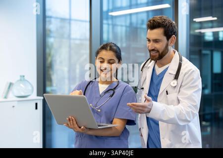 Un medico e un'infermiera rivedono felicemente un portatile insieme in un ambiente medico moderno, sorridendo. Entrambi indossano stetoscopi e abbigliamento medico. Foto Stock