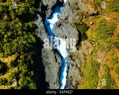Vista aerea della cascata hraunfossar che si tuffa attraverso la vegetazione lussureggiante e il terreno roccioso di reykholt, islanda. Foto Stock