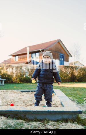 Un ragazzo allegro in abiti caldi invernali giocherà gioiosamente in una scatola di sabbia piena di giocattoli colorati, con una casa accogliente come sfondo delizioso per il suo outdoo Foto Stock