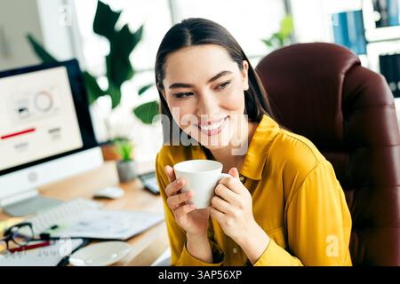 Donna professionista in un ufficio moderno sorridente e in mano una tazza di caffè, godendo momenti di relax mentre lavora al computer Foto Stock