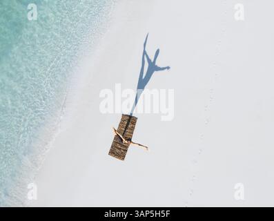 Vista aerea di una spiaggia tranquilla con una donna che pratica yoga su un tappeto, circondata da acque turchesi cristalline e dolci onde, Thoddoo, Maldive. Foto Stock