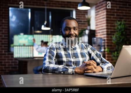 Ritratto di un uomo afroamericano sorridente e che guarda la macchina fotografica, seduto a una scrivania con un portatile. L'ufficio è poco illuminato con una parete in mattoni, ideale per i turni di notte e il monitoraggio del mercato azionario. Foto Stock